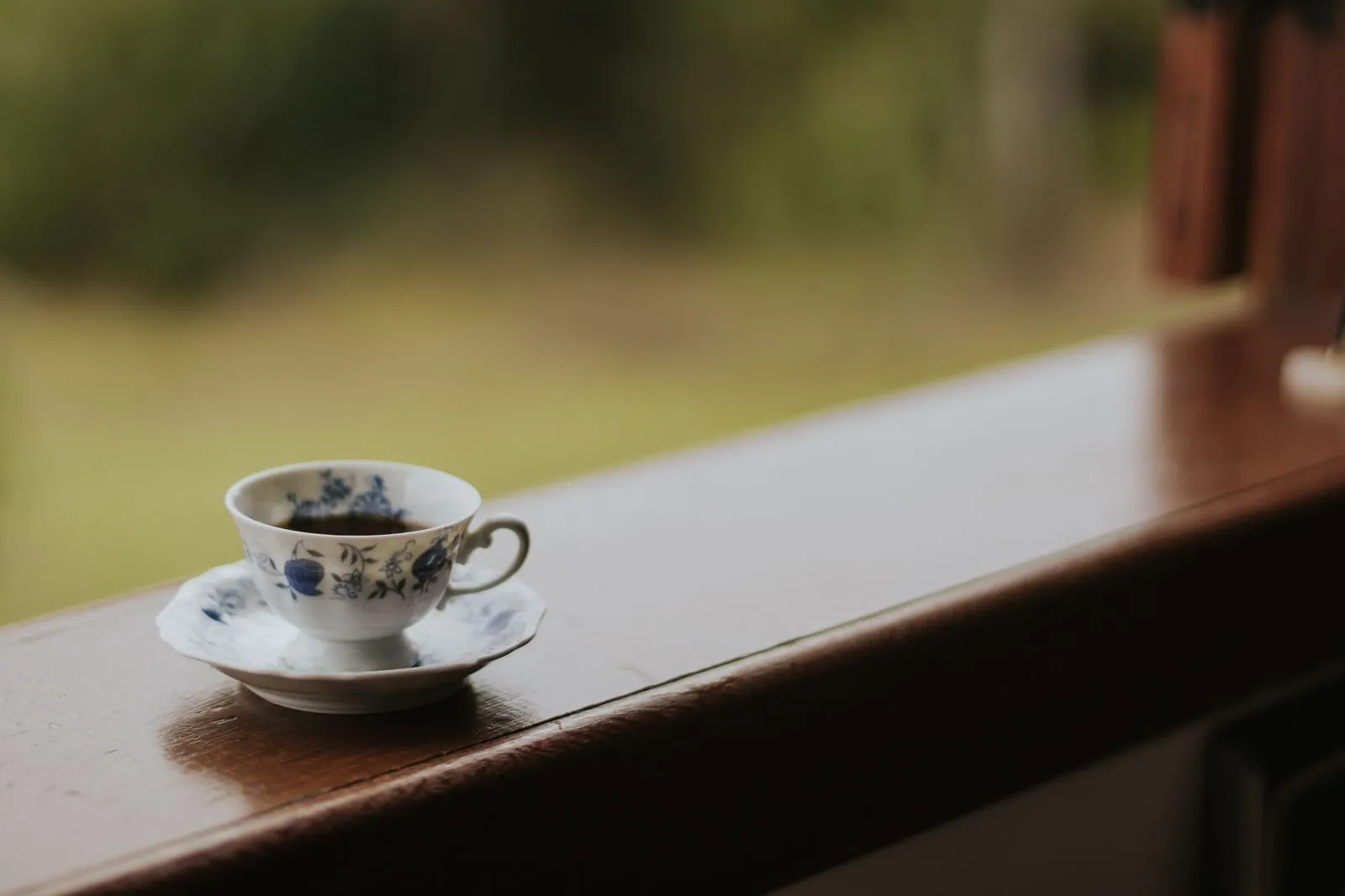 木頭窗台上放著一只藍花瓷茶杯，窗外是模糊的綠色草地